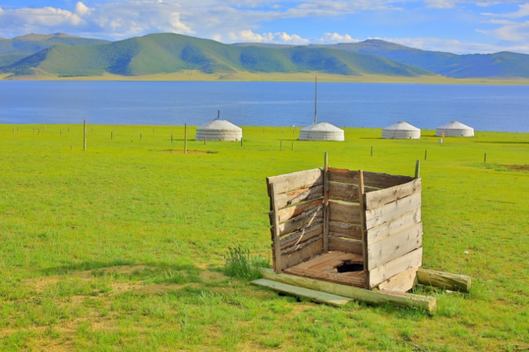 草原の中に低い板で囲まれたトイレと、遠くに湖とモンゴル式の移動住居のゲル