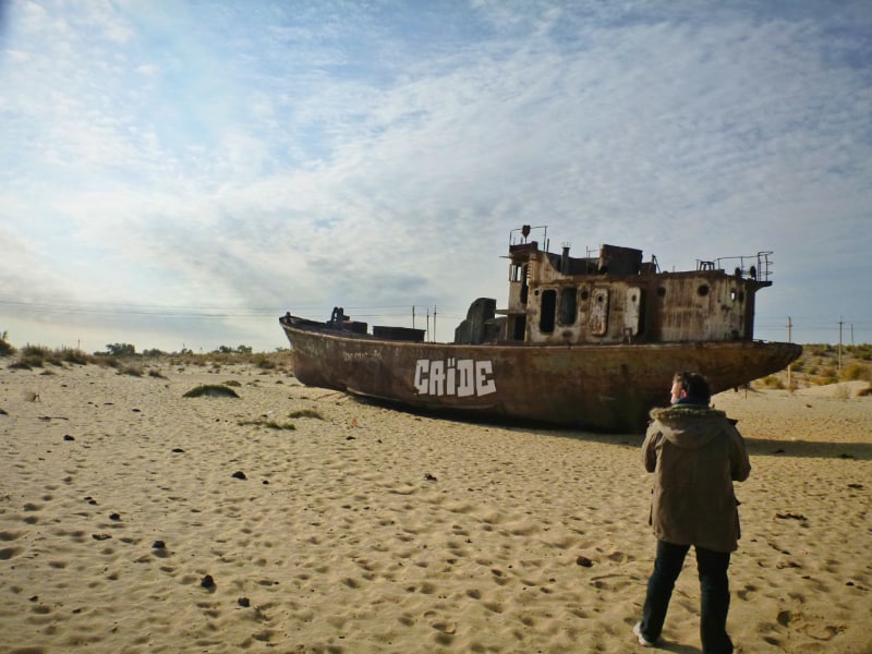 砂地にさび付いた船の残骸。手前に男性が後ろ向きで立っている写真