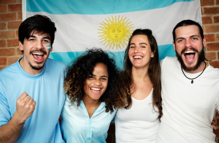 ラテン系？の男性二人、女性二人がアルゼンチン国旗の前で肩を組んでいる