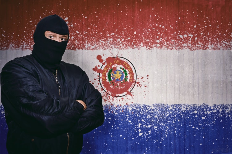 パラグアイ国旗の前に覆面の男が腕を組んで立っている