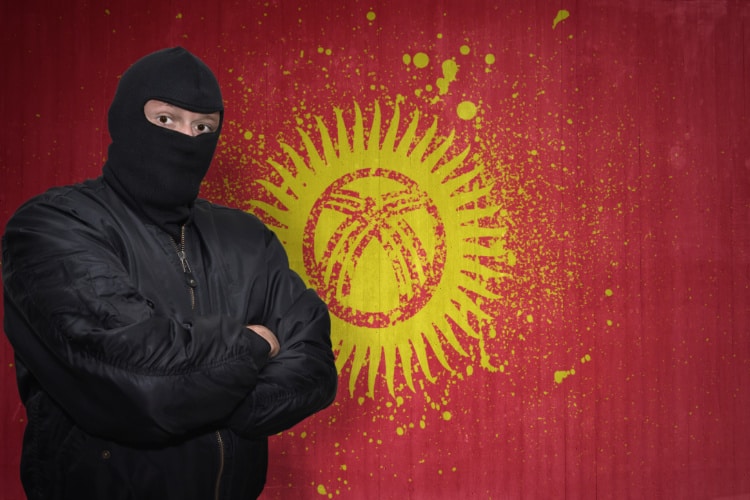 キルギス国旗の前にマスクの男が腕を組んで立っている
